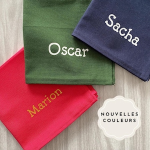 Può includere: Tre tovagliette da tavola in tessuto con nomi ricamati: Marion in oro su rosso, Oscar in bianco su verde e Sacha in bianco su blu navy. Le tovagliette sono impilate con il testo "NOUVELLES COULEURS" visibile in un cerchio bianco.