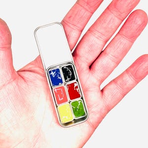 May include: A small metal tin containing six watercolour paint pans. The pans are filled with blue, black, red, pink, yellow, and green paint.