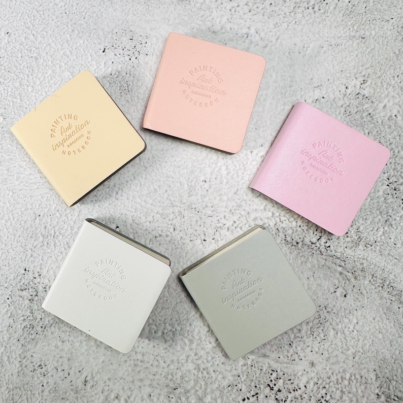 May include: Five square notebooks in pastel colors including yellow, light pink, bright pink, white, and light gray. Each notebook features a debossed circular logo on the front cover with the text Painting Art inspiration NOTEBOOK.