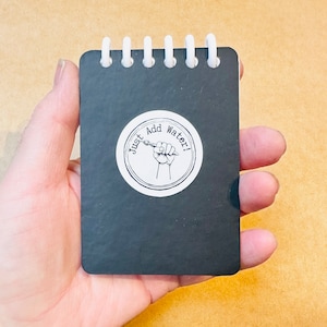 May include: A small black spiral-bound notebook featuring a white circular sticker on the front cover. The sticker displays a black and white illustration of a hand holding a paintbrush with the text Just Add Water! printed in a circular arc.