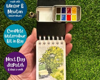 May include: A portable watercolor kit featuring a small black spiral-bound sketchbook with a landscape painting of a tree on the cover. Attached to the top is a metal tin containing eight small rectangular pans of Winsor & Newton watercolors.