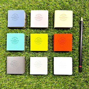 May include: Eight square notebooks in various colors including blue, pink, yellow, teal, lime, orange, gray, and white, each embossed with the text Painting Art Inspiration Notebook, arranged in a grid next to a black pencil.