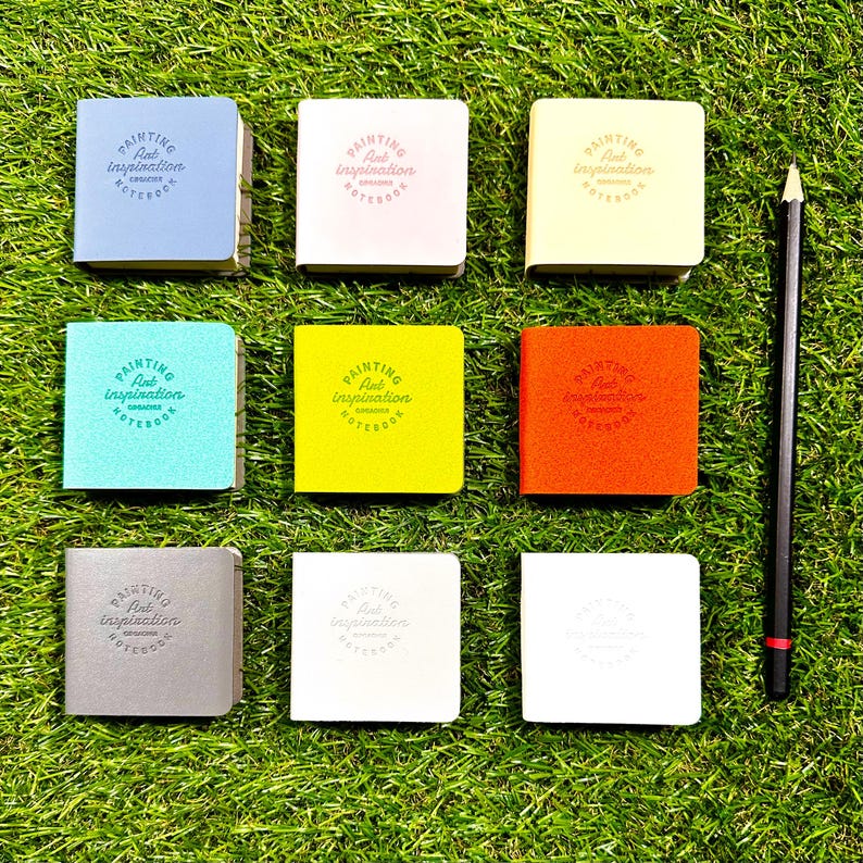 May include: Eight square notebooks in various colors including blue, pink, yellow, teal, lime, orange, gray, and white, each embossed with the text Painting Art Inspiration Notebook, arranged in a grid next to a black pencil.