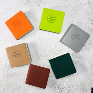 May include: Six square notebooks in orange, lime green, gray, tan, dark green, and brown. Each notebook features a circular embossed logo on the front cover with the text "PAINTING Art inspiration NOTEBOOK" arranged in a circular pattern.