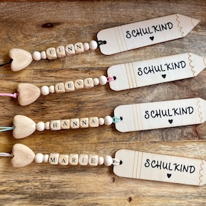 Könnte beinhalten: Vier Holz-Bleistifte-Lesezeichen mit dem Text "Schulkind" und einem kleinen Herz, jedes an einer Holzperlenkette mit einem Herz-Anhänger und Buchstabenperlen, die die Namen Finn, Levi, Hanna und Marie buchstabieren.