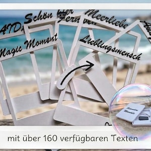 Könnte beinhalten: Eine Sammlung weißer, rechteckiger, rahmenförmiger Holzausschnitte mit verschiedenen schwarz gedruckten Texten wie Magic Moment und Lieblingsmensch. Eine kleine kreisförmige Blase zeigt eine weiße Geschenkbox mit einem silbernen Armband.