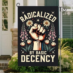 Op de afbeelding: Een zwarte tuinvaandel met de tekst "RADICALIZED BY BASIC DECENCY" in wit en geel. De vlag toont een vuist, bloemen en sterren in een kleurrijk ontwerp. De vlag hangt aan een zwarte metalen standaard.