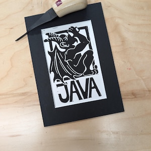 May include: A black and white linocut print of a devil-like creature with wings, sitting and holding a glass. The print is framed in a black mat and has the word "JAVA" printed below the image.