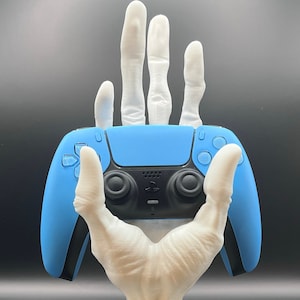 May include: A white, hand-shaped gaming controller stand holding a blue and black controller. The stand has a detailed, textured design and a circular base. The controller is a modern design with black buttons and a blue face.