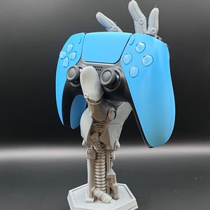 May include: A light blue and black video game controller held by a gray robotic arm stand. The stand has a hexagonal base and a spring-like design in the center. The controller has buttons and joysticks.