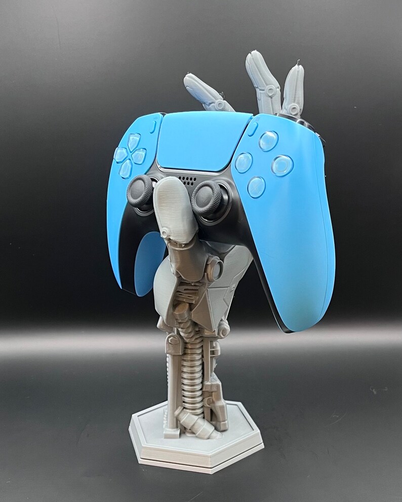 May include: A light blue and black video game controller held by a gray robotic arm stand. The stand has a hexagonal base and a spring-like design in the center. The controller has buttons and joysticks.