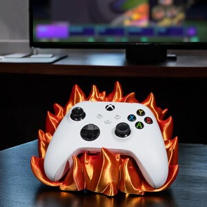 May include: A white Xbox controller rests in a 3D-printed orange and gold flame-shaped holder. The controller has black buttons and colored action buttons. The holder is on a dark surface, with a TV in the background.