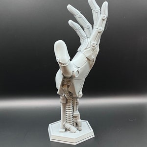 May include: A light gray, 3D-printed robotic hand sculpture. The hand is posed with fingers extended, showcasing intricate mechanical details. It is mounted on a hexagonal base, with visible springs and joints.