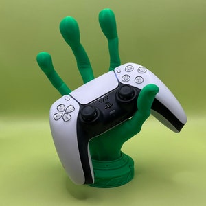 May include: A green, hand-shaped controller stand holding a white and black game controller. The stand has a detailed base and the controller features black joysticks and buttons. The background is a solid light green.