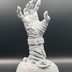 May include: A detailed, gray sculpture of a reaching hand, with visible skin texture and a band around the wrist. The hand emerges from a base with a textured surface, suggesting a zombie or monster theme.