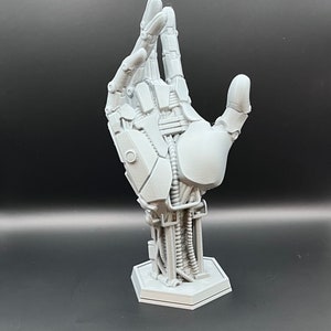May include: A gray, 3D-printed robotic hand sculpture. The hand is posed with the thumb and index finger extended, showcasing intricate mechanical details. It is mounted on a hexagonal base, perfect for display.