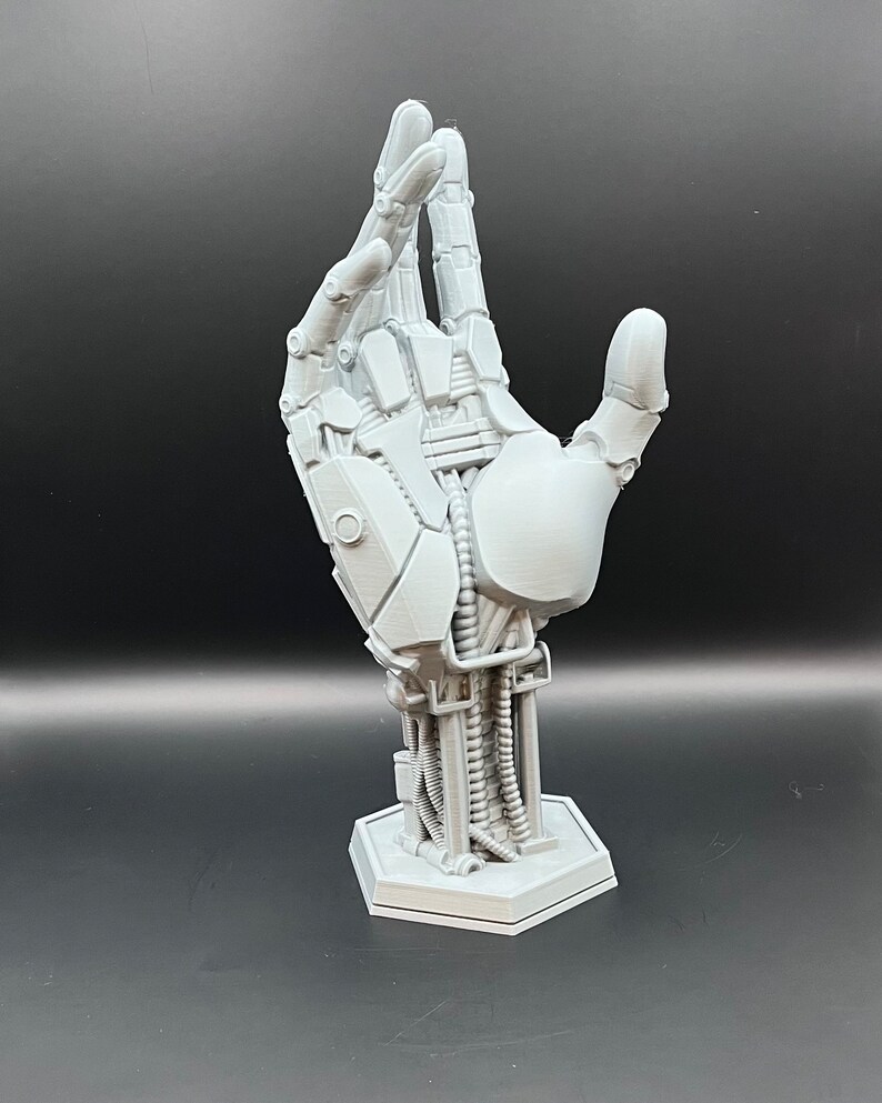May include: A gray, 3D-printed robotic hand sculpture. The hand is posed with the thumb and index finger extended, showcasing intricate mechanical details. It is mounted on a hexagonal base, perfect for display.