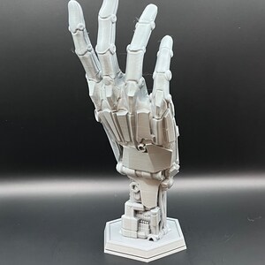 May include: A gray, articulated robotic hand sculpture, with detailed mechanical joints and a hexagonal base. The hand is posed in a gesture, showcasing its intricate design and construction. The sculpture is made of a light gray material.