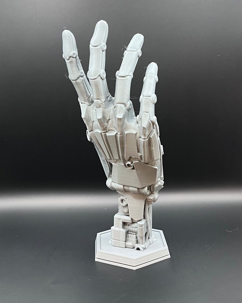 May include: A gray, articulated robotic hand sculpture, with detailed mechanical joints and a hexagonal base. The hand is posed in a gesture, showcasing its intricate design and construction. The sculpture is made of a light gray material.