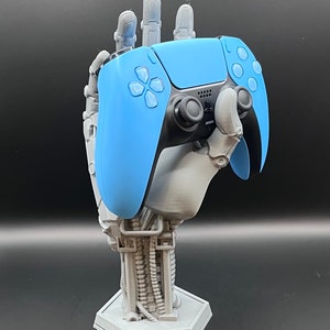 May include: A light blue and black video game controller rests in a gray, robotic hand-shaped stand. The stand has a hexagonal base and detailed mechanical arm design. The controller's buttons and joysticks are visible, set against a dark background.