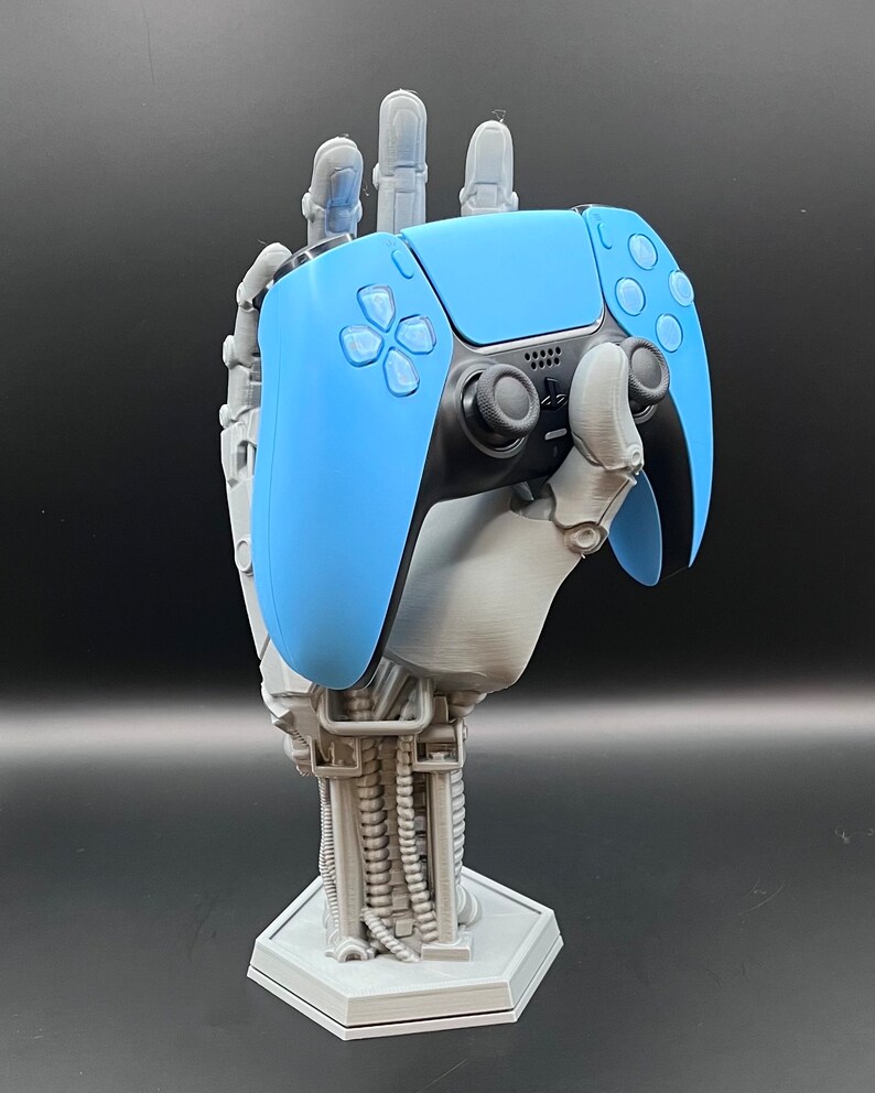 May include: A light blue and black video game controller rests in a gray, robotic hand-shaped stand. The stand has a hexagonal base and detailed mechanical arm design. The controller's buttons and joysticks are visible, set against a dark background.