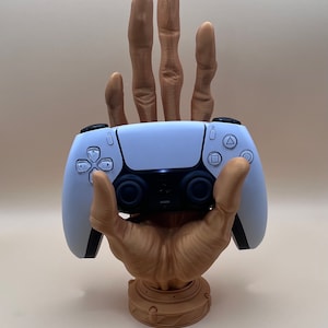 May include: A brown, hand-shaped gaming controller stand holding a white and black controller. The stand has a detailed design, with fingers extending upwards. The controller has buttons and a touchpad.