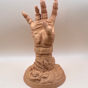 May include: A tan-colored, detailed sculpture of a hand reaching upwards. The hand is attached to a wrist with a belt and buckle. The base features a cockroach and other sculpted details. The sculpture is likely a decorative item.