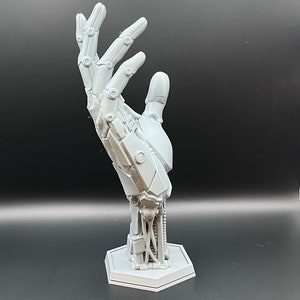 May include: A gray, 3D-printed robotic hand sculpture. The hand is posed in an open gesture, showcasing intricate mechanical details. It is mounted on a hexagonal base, with visible wiring and joint structures, suggesting a futuristic or technological theme.