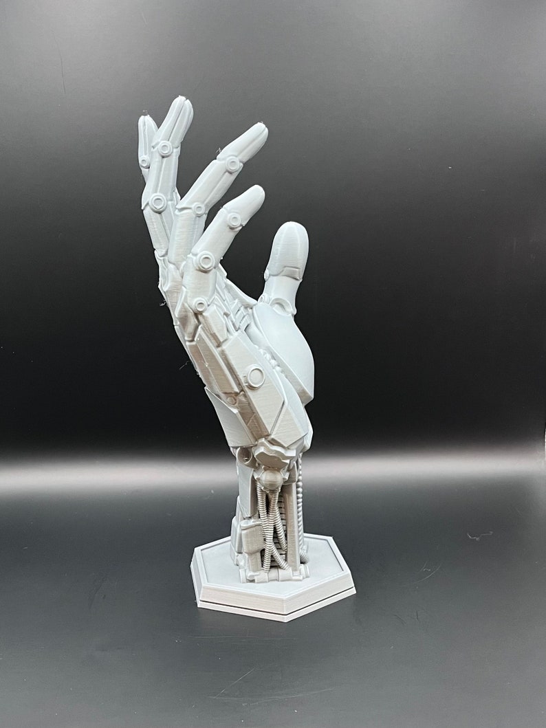 May include: A gray, 3D-printed robotic hand sculpture. The hand is posed in an open gesture, showcasing intricate mechanical details. It is mounted on a hexagonal base, with visible wiring and joint structures, suggesting a futuristic or technological theme.