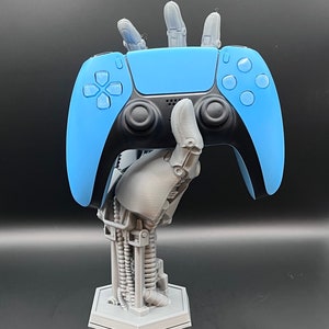 May include: A light blue and black video game controller held by a gray robotic hand. The hand is attached to a mechanical arm and base, creating a unique display stand for the controller. The stand is gray and has a hexagonal base.