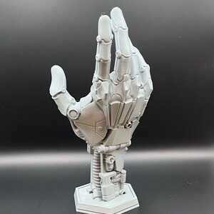 May include: A gray, 3D-printed robotic hand sculpture. The hand is posed in a gesture, with detailed mechanical components visible. It is mounted on a hexagonal base, showcasing a futuristic aesthetic. The sculpture is a solid, monochromatic piece.