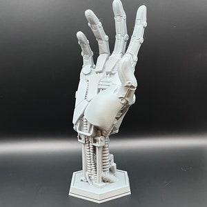 May include: A light gray, 3D-printed robotic hand sculpture. The hand is in an open position, revealing intricate mechanical details. It is mounted on a hexagonal base. The sculpture is made of plastic and is a futuristic art piece.