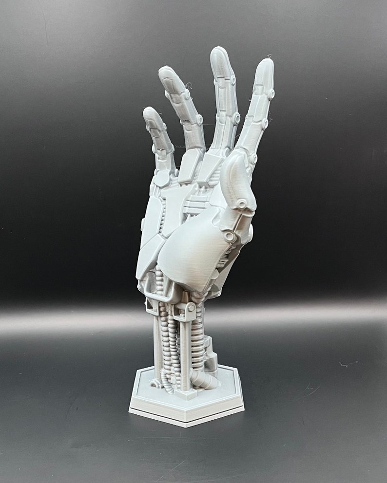 May include: A light gray, 3D-printed robotic hand sculpture. The hand is in an open position, revealing intricate mechanical details. It is mounted on a hexagonal base. The sculpture is made of plastic and is a futuristic art piece.