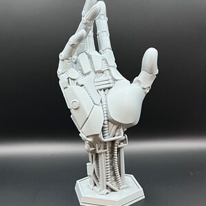 May include: A gray, 3D-printed robotic hand sculpture. The hand is posed with the fingers slightly curved, revealing intricate mechanical details, including joints, wires, and a hexagonal base. The sculpture is a futuristic art piece.