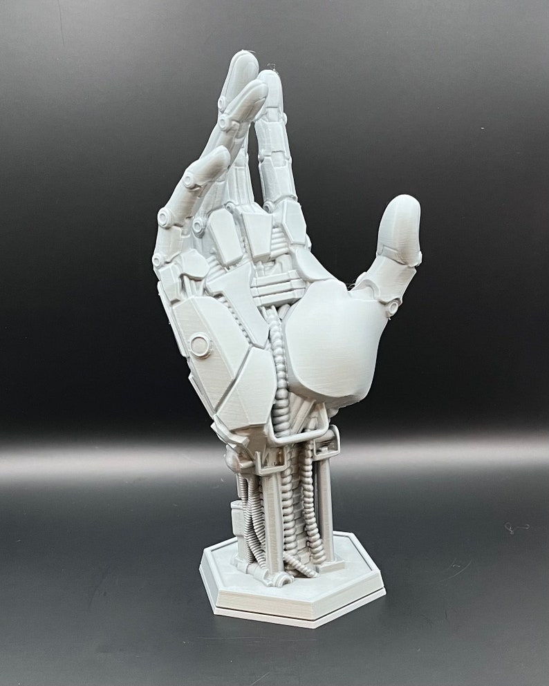 May include: A gray, 3D-printed robotic hand sculpture. The hand is posed with the fingers slightly curved, revealing intricate mechanical details, including joints, wires, and a hexagonal base. The sculpture is a futuristic art piece.
