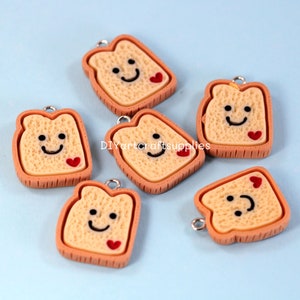 May include: Six brown and beige resin charms shaped like slices of toast with a smiling face and a red heart on each. The charms have a hole at the top for hanging.