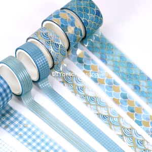 May include: A set of six blue and gold washi tapes with various patterns, including scales, stripes, and gingham. The tapes are rolled up and arranged in a fan shape.