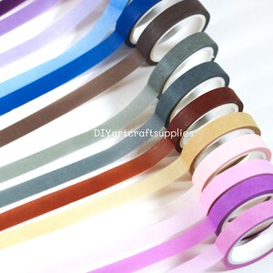 May include: A set of 12 washi tapes in various colors, including purple, blue, brown, gray, beige, pink, and red. The tapes are arranged in a fan shape, with the ends of the tapes overlapping. The tapes are all 15mm wide and are made of high-quality paper.