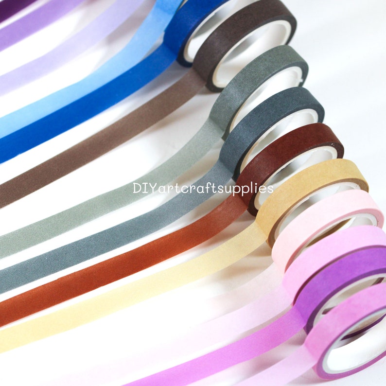 May include: A set of 12 washi tapes in various colors, including purple, blue, brown, gray, beige, pink, and red. The tapes are arranged in a fan shape, with the ends of the tapes overlapping. The tapes are all 15mm wide and are made of high-quality paper.