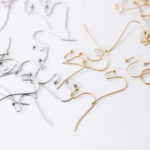 May include: A close-up of a collection of silver and gold earring hooks. The hooks are made of metal and have a small ball at the end. The hooks are arranged in a random pattern on a white surface.