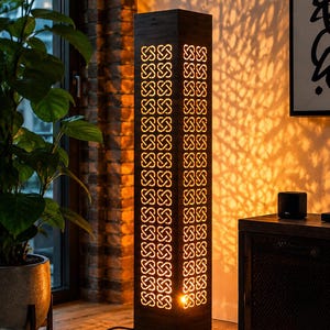 Può includere: Una lampada da terra alta e rettangolare in legno scuro con un motivo ritagliato ripetitivo di nodi intrecciati. La lampada emette una luce calda e dorata attraverso le aperture, proiettando motivi complessi simili ad ombre sulla parete.