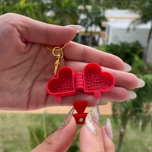 May include: A red heart-shaped keychain with a gold chain and a small red heart charm. The keychain is open to reveal a delicate, intricate design inside.