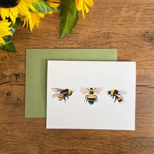 May include: A white card with three watercolor illustrations of bumblebees. The bumblebees are yellow and black with translucent wings. The card is on a green cardstock background.