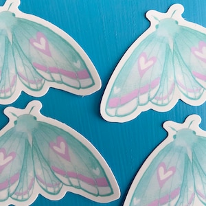 May include: Four teal blue and pink moth stickers with heart details. The stickers are on a blue background.