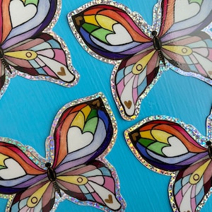 May include: Four rainbow butterfly stickers with a silver glitter border. Each butterfly has a rainbow wing pattern with white hearts and a gold outline.