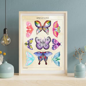May include: A framed print featuring nine colorful butterfly illustrations. The butterflies are in various colors, including pink, blue, purple, and rainbow hues. The text "SPREAD LOVE" is at the top. The artwork is displayed on a light beige background.
