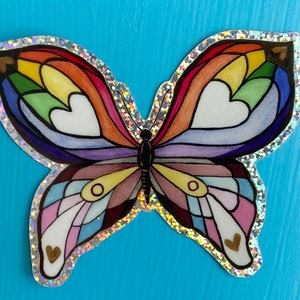May include: A rainbow-colored butterfly sticker with a glittery border and gold hearts. The butterfly's wings are made up of different colors of the rainbow, and the hearts are scattered throughout the wings.