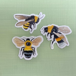 May include: Three bee stickers with black and yellow stripes and translucent wings. The bees are in different poses, with one flying, one facing forward, and one facing to the side.
