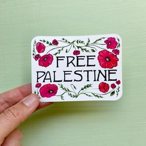 May include: A white sticker with the words "FREE PALESTINE" in black, surrounded by red poppy flowers and green leaves. The sticker is rectangular with rounded corners and is held by a hand against a light green background.