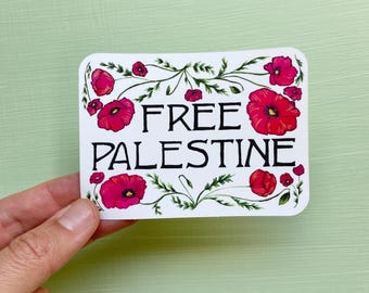 Può includere: Un adesivo bianco con la scritta "FREE PALESTINE" in nero, circondata da fiori di papavero rossi e foglie verdi. L'adesivo rettangolare con angoli arrotondati è tenuto in mano su uno sfondo verde chiaro.